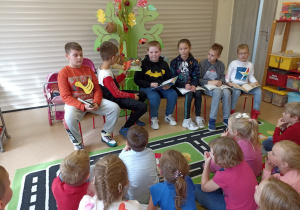 Dzieci ze Szkoły Podstawowej Nr 2 z Oddziałami Integracyjnymi w Ozorkowie czytają przedszkolakom bajki siedząc przed nimi na krzesłach
