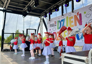Występ dziewczynek na scenie- są ubrane w czerwone koszulki, szare spódniczki, mają na głowie opaski- kokardki, a w rękach trzymają kolorowe pompony