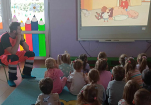 Ratownik medyczny wraz z dziećmi ogląda film edukacyjny na tablicy multimedialnej na temat zasad udzielania pierwszej pomocy