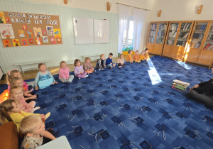 Dzieci siedzą na dywanie w bibliotece, a przed nimi siedzi pani.