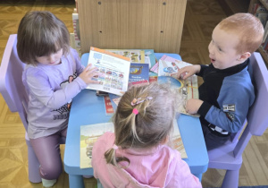 Troje dzieci siedzi przy stole i przegląda książki.