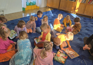 Dzieci siedzą swobodnie na dywanie i trzymają w rękach książki. Obok nich siedzi pani, która patrzy na dzieci.