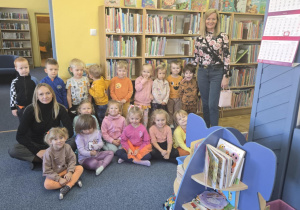 Dzieci z grupy Misie wraz z paniami pozują do zdjęcia na tle książek. Część dzieci siedzi, pozostałe stoją. Jedna z Pań siedzi obok dzieci, a druga stoi razem z dziećmi.