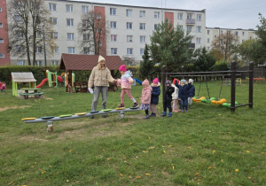 Dzieci ustawione w rzędzie przechodzą po równoważni, przeliczają jej elementy, a nauczycielka asekuruje je podczas ćwiczenia.