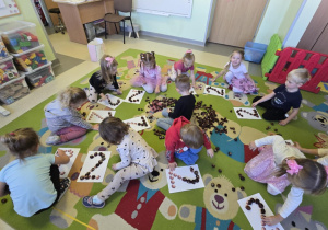 Grupa dzieci siedzi na dywanie, każde z nich ma przed sobą kartę pracy z cyfrą 2, na dywanie rozrzucone są kasztany, które dzieci układają na karcie w odpowiednim kierunku, zgodnie z tym jak pisze się cyfrę dwa.