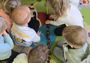 Na dywanie z taśmy malarskiej przyklejona jest cyfra jeden, a grupa dzieci układa na niej kasztany.