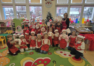 Dzieci ubrane w czerwone fartuszki i czapki mikołajkowe pozują do zdjęcia wraz z panią Marzenką