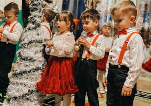 Dzieci stoją w szeregu obok choinki. Jeden z chłopców recytuje wierszyk do mikrofonu.