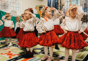 Dziewczynki tańczą na scenie, zwrócone w tę samą stronę, z dłonią uniesioną nad głową.