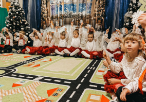 Dzieci, odświętnie ubrane, siedzą na tle świątecznej dekoracji i trzymają w dłoniach śnieżynki.