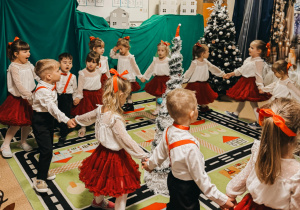 Dzieci tańczą w kole, w środku stoi choinka.