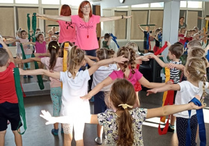 Dzieci stoją prosto z rozłożonymi na bok rękami i ćwiczą z panią Kamilą w sali gimnastycznej.