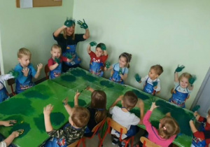 Dzieci i nauczycielka pokazują zielony kolor na rękach po zmieszaniu w rytm muzyki niebieskiego i żółtego koloru