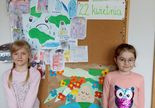 Dwie dziewczynki pozują do zdjęcia przy tablicy ze wspólną pracą plastyczną