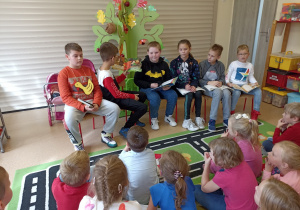 Dzieci ze Szkoły Podstawowej Nr 2 z Oddziałami Integracyjnymi w Ozorkowie czytają przedszkolakom bajki siedząc przed nimi na krzesłach