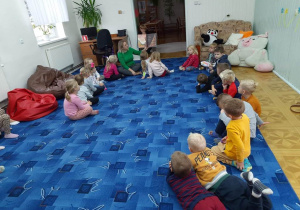 dzieci siedzą w kole na dywanie i słuchają opowiadania czytanego przez panią bibliotekarkę