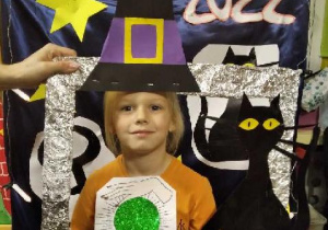 Dziewczynka pozuje na tle dekoracji andrzejkowej do zdjęcia w ramce z kapeluszem czarodziejki na głowie..