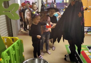 Nauczycielka przebrana za wróżkę podaje dzieciom monety do wróżby z miską wody..