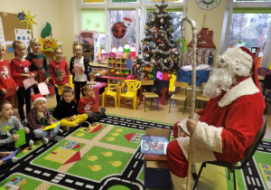 Mikołaj siedzi na krześl obok ma prezenty, a przed nim na dywanie siedzą dzieci.