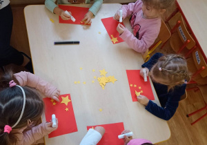 Dzieci tworzą flagę Chin przyklejając gotowe elementy na czerwoną kartkę papieru według wzoru