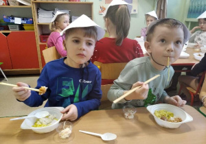 Chłopcy siedzą przy stoliku i jedzą pałeczkami w białych czapkach z kartonu.