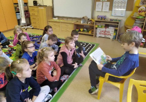 Chłopiec na krzesełku czyta bajkę dzieciom siedzącym przed nim.
