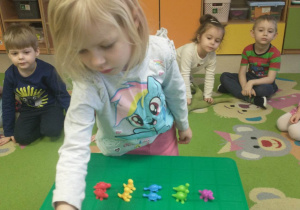 dziewczynka stoi na dywanie, dziewczynka układa sekwencje- kolorowe dinozaury