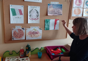 Nauczycielka opowiada dzieciom o symbolach narodowych Włoch, pokazuje jak w prawidłowy sposób namalować farbami flagę Włoch