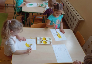 Dzieci przy stoliku wypełniają żółtą farbą kontury obrazka przedstawiającego kontener na plastik, a zieloną kontener na szkło