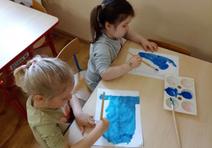 Dzieci przy stoliku wypełniają niebieską farbą kontury obrazka przedstawiającego kontener na papier