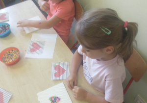 dziewczynki siedzą przy stoliku, układają koraliki według szablonu