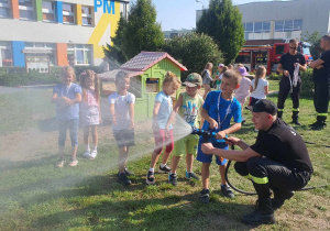 Dzieci kolejno trzymają w rękach wąż strażacki i leją wodą