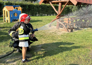 Dzieci kolejno trzymają w rękach wąż strażacki i leją wodą