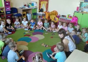 Dzieci siedzą w kole na dywanie z Panią na zajęciach z języka angielskiego.