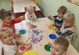 Uśmiechnięte dzieci siedzą przy stoliku i nawlekają pocięte kolorowe słomki na wykałaczki przymocowane do plastikowych talerzyków