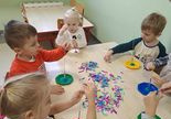 Dzieci siedzą przy stoliku i nawlekają krótkie kolorowe rurki na wykałaczki.