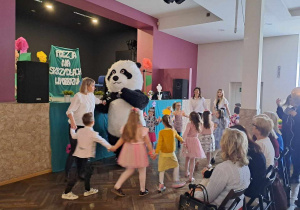 Dzieci wraz z nauczycielkami tańczą przed sceną z Misiem Pandą